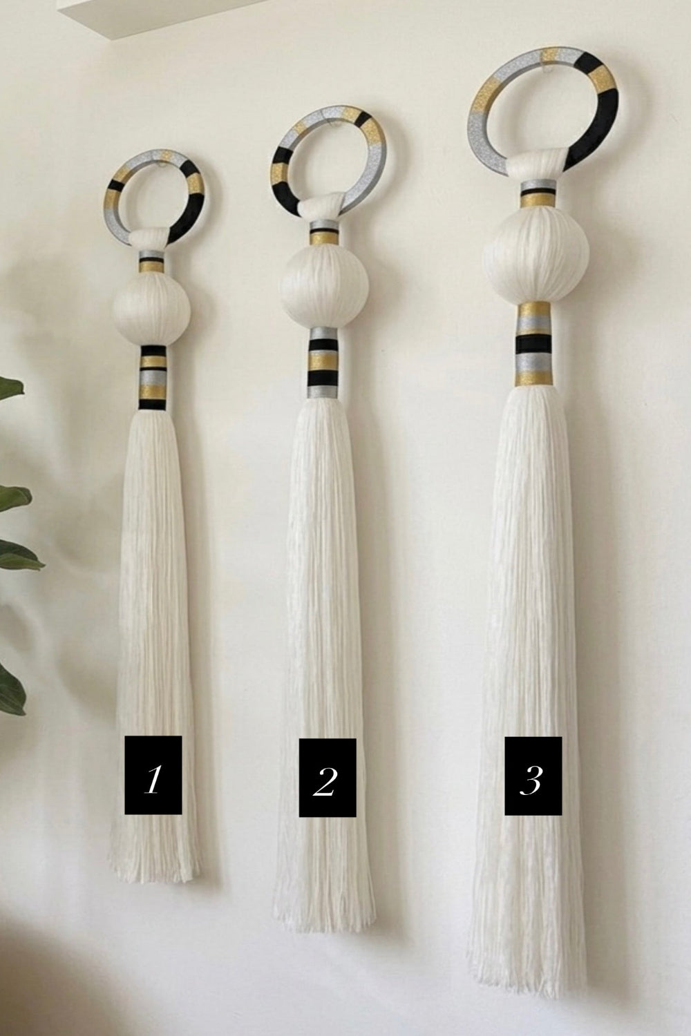 Three decorative wall-mounted tassel ornaments are shown hanging on a light wall, each with a circular top, a white spherical section, a striped black-and-gold neck, and a long white tassel. Black labels at the bottom read 1, 2, and 3.
