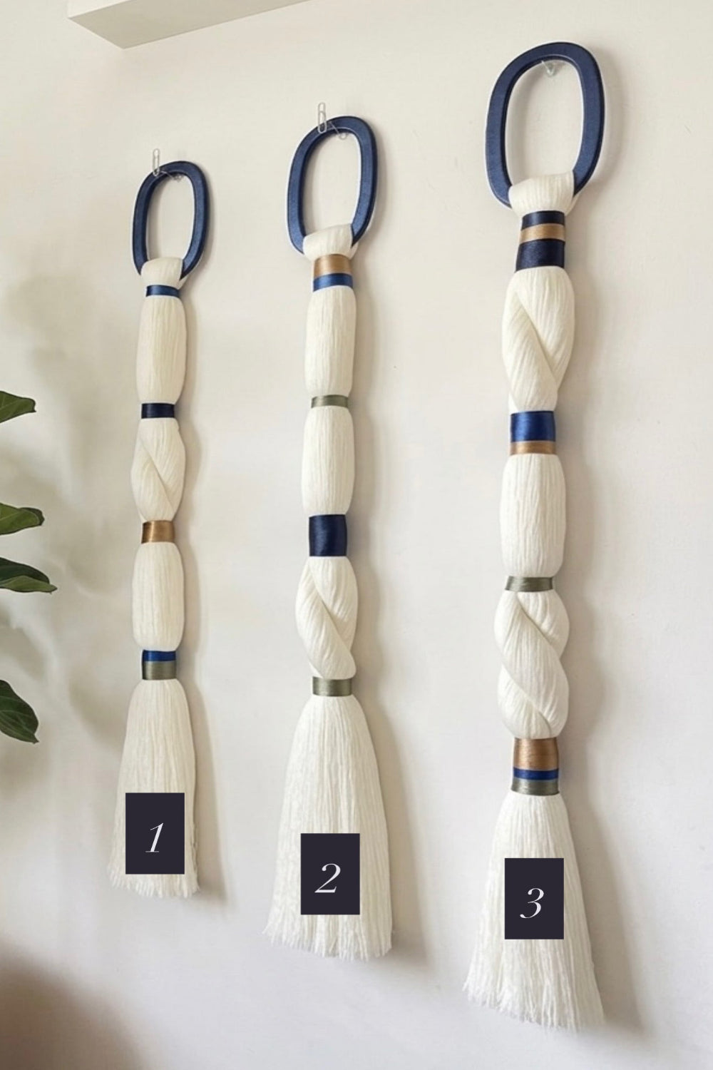Three decorative wall-mounted rope ornaments or tassels displayed vertically on a white wall, labeled 1, 2, and 3 with black tags. Each has a blue loop at the top, cream and white rope sections with blue and metallic bands, and a frayed white end.
