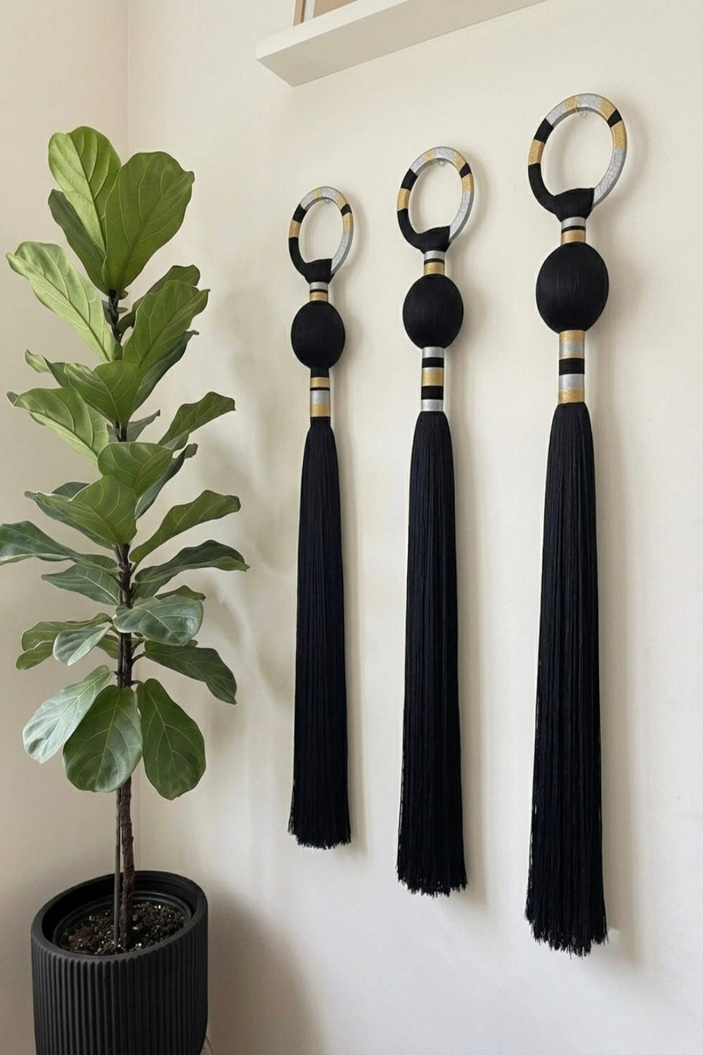 Three black decorative wall tassels with circular ring tops and gold-and-white striped accents, displayed on a white wall next to a potted fiddle-leaf fig plant.