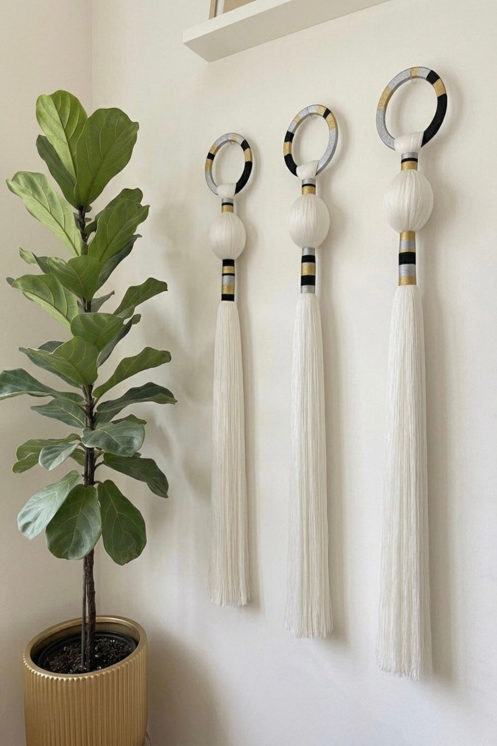 Three decorative wall-mounted tassel ornaments with circular ring tops are displayed on a white wall beside a potted fiddle-leaf fig plant. The ornaments are primarily off-white/cream with black and metallic gold accents near the rings and tassels.