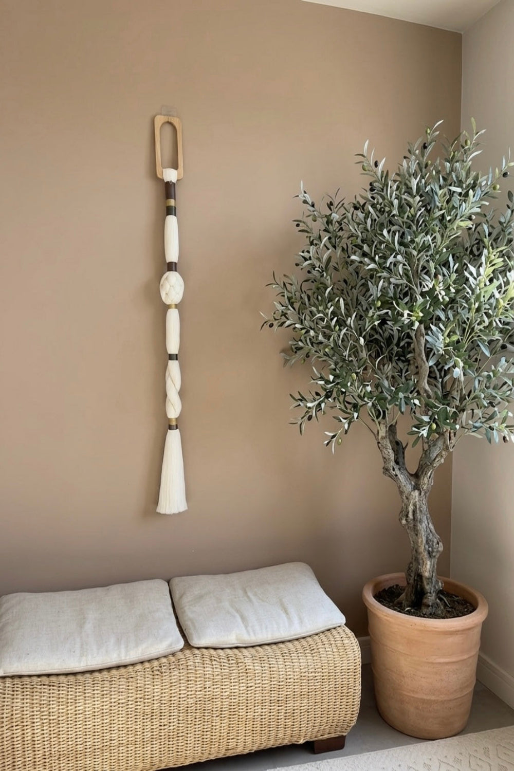 A bohemian-style woven wall hanging with a wooden top frame, cream beads, and a tassel, displayed on a taupe wall above a wicker bench next to a potted olive tree.