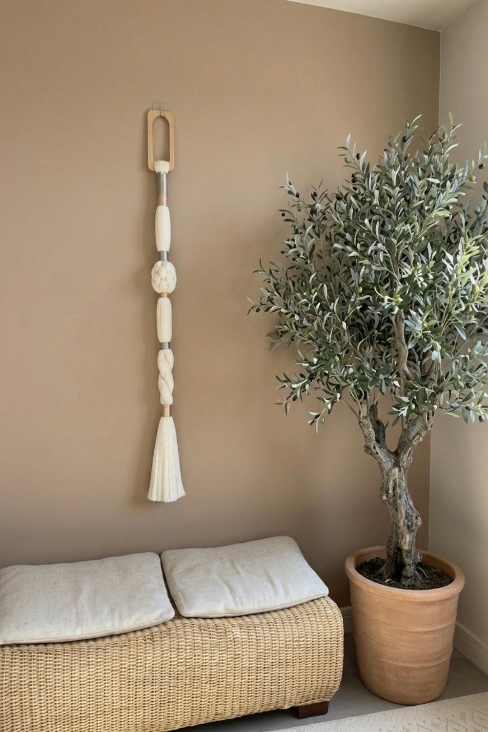 A bohemian-style cream macrame wall hanging with a wooden ring and tassel, displayed on a warm taupe wall above a woven rattan bench with white cushions, next to a potted olive tree.