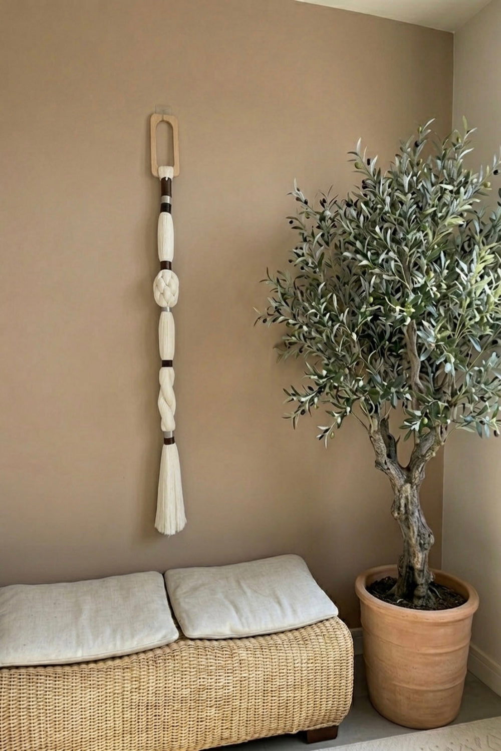 A cream macrame wall hanging with a wooden hanger is displayed on a warm beige wall in a styled room corner next to a potted olive tree and a woven bench with cushions.