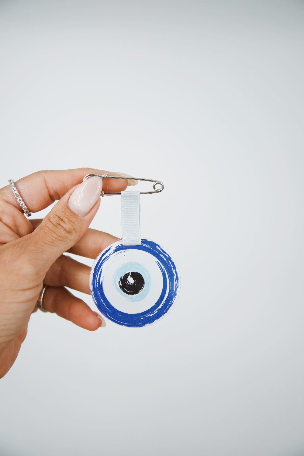 A hand holding a silver safety pin bag clip with a blue and white evil eye charm against a light gray background.